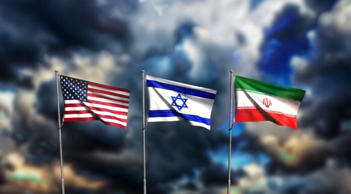 Iran-Krieg: Das wird nun teurer Die Flaggen von den USA, Isreal und Iran wehen vor einem dunklen, wolkenverhangenen Himmel.