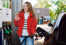 So sparen Sie jetzt beim Tanken bares Geld Ein Porträt einer jungen Frau, die mit einem Telefon in der Hand an einer Tankstelle neben ihrem Auto steht, das gerade getankt wird.