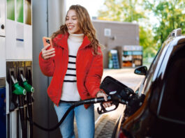 So sparen Sie jetzt beim Tanken bares Geld Ein Porträt einer jungen Frau, die mit einem Telefon in der Hand an einer Tankstelle neben ihrem Auto steht, das gerade getankt wird.
