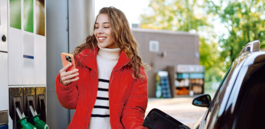 So sparen Sie jetzt beim Tanken bares Geld Ein Porträt einer jungen Frau, die mit einem Telefon in der Hand an einer Tankstelle neben ihrem Auto steht, das gerade getankt wird.