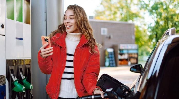So sparen Sie jetzt beim Tanken bares Geld Ein Porträt einer jungen Frau, die mit einem Telefon in der Hand an einer Tankstelle neben ihrem Auto steht, das gerade getankt wird.