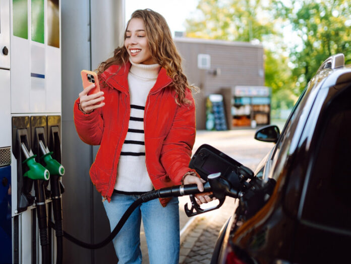 Ein Porträt einer jungen Frau, die mit einem Telefon in der Hand an einer Tankstelle neben ihrem Auto steht, das gerade getankt wird.