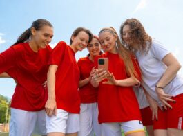 Kita, Schule, Sport: Gruppenkasse einfach organisieren Eine Gruppe junger Fußballerinnen in roten und weißen Trikots lächelt, während sie im Freien auf das Telefon in der Hand einer der Frauen schauen.