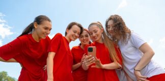 Kita, Schule, Sport: Gruppenkasse einfach organisieren Eine Gruppe junger Fußballerinnen in roten und weißen Trikots lächelt, während sie im Freien auf das Telefon in der Hand einer der Frauen schauen.
