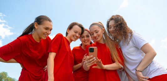 Kita, Schule, Sport: Gruppenkasse einfach organisieren Eine Gruppe junger Fußballerinnen in roten und weißen Trikots lächelt, während sie im Freien auf das Telefon in der Hand einer der Frauen schauen.