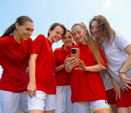 Kita, Schule, Sport: Gruppenkasse einfach organisieren Eine Gruppe junger Fußballerinnen in roten und weißen Trikots lächelt, während sie im Freien auf das Telefon in der Hand einer der Frauen schauen.