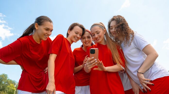 Kita, Schule, Sport: Gruppenkasse einfach organisieren Eine Gruppe junger Fußballerinnen in roten und weißen Trikots lächelt, während sie im Freien auf das Telefon in der Hand einer der Frauen schauen.