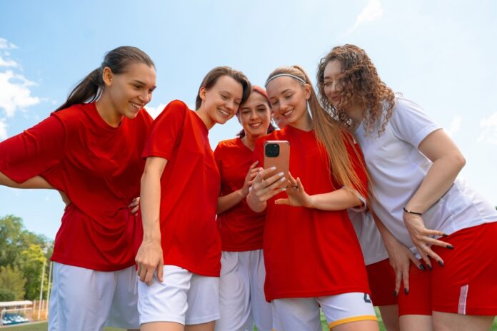Eine Gruppe junger Fußballerinnen in roten und weißen Trikots lächelt, während sie im Freien auf das Telefon in der Hand einer der Frauen schauen.