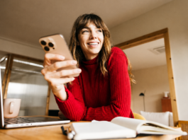 Einfach. Sicher. Online: Kartenzahlung mit 3-D Secure Eine lächelnde Frau im roten Pullover sitzt mit Smartphone, Laptop und Notizbuch an einem Tisch im Homeoffice.