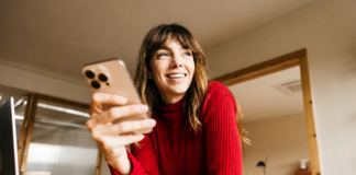 Einfach. Sicher. Online: Kartenzahlung mit 3-D Secure Eine lächelnde Frau im roten Pullover sitzt mit Smartphone, Laptop und Notizbuch an einem Tisch im Homeoffice.