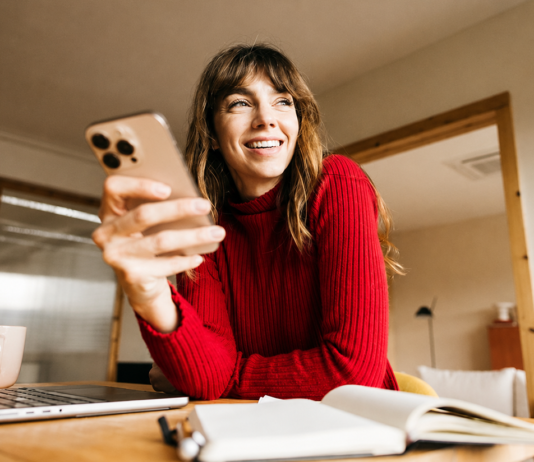 Einfach. Sicher. Online: Kartenzahlung mit 3-D Secure Eine lächelnde Frau im roten Pullover sitzt mit Smartphone, Laptop und Notizbuch an einem Tisch im Homeoffice.
