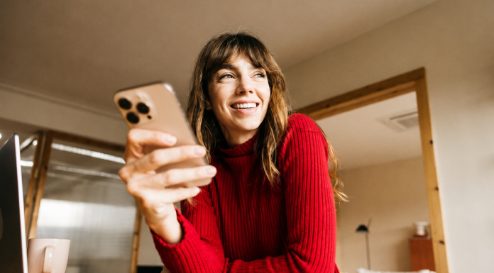 Einfach. Sicher. Online: Kartenzahlung mit 3-D Secure Eine lächelnde Frau im roten Pullover sitzt mit Smartphone, Laptop und Notizbuch an einem Tisch im Homeoffice.
