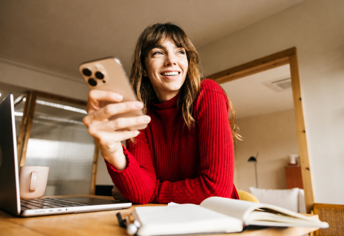 Eine lächelnde Frau im roten Pullover sitzt mit Smartphone, Laptop und Notizbuch an einem Tisch im Homeoffice.