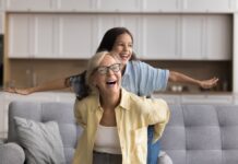 Wie funktioniert die Frühstartrente? Oma und Enkelin sitzen auf einem Sofa und lachen, die Enkelin spreizt die Arme wie bei einem Flugzeug, Oma hält sie fest