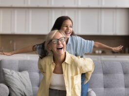 Wie funktioniert die Frühstartrente? Oma und Enkelin sitzen auf einem Sofa und lachen, die Enkelin spreizt die Arme wie bei einem Flugzeug, Oma hält sie fest