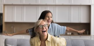 Wie funktioniert die Frühstartrente? Oma und Enkelin sitzen auf einem Sofa und lachen, die Enkelin spreizt die Arme wie bei einem Flugzeug, Oma hält sie fest
