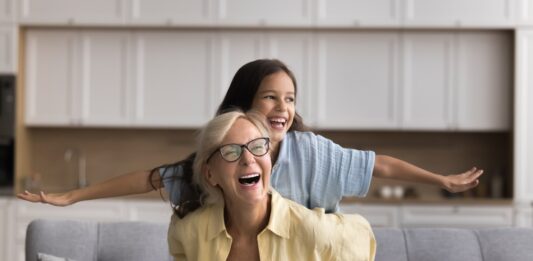 Wie funktioniert die Frühstartrente? Oma und Enkelin sitzen auf einem Sofa und lachen, die Enkelin spreizt die Arme wie bei einem Flugzeug, Oma hält sie fest