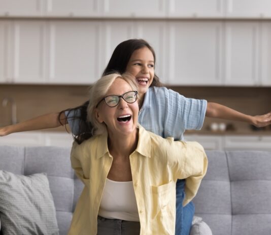 Wie funktioniert die Frühstartrente? Oma und Enkelin sitzen auf einem Sofa und lachen, die Enkelin spreizt die Arme wie bei einem Flugzeug, Oma hält sie fest