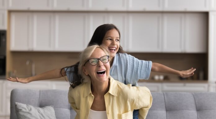 Wie funktioniert die Frühstartrente? Oma und Enkelin sitzen auf einem Sofa und lachen, die Enkelin spreizt die Arme wie bei einem Flugzeug, Oma hält sie fest