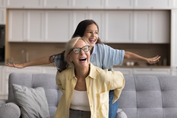 Oma und Enkelin sitzen auf einem Sofa und lachen, die Enkelin spreizt die Arme wie bei einem Flugzeug, Oma hält sie fest